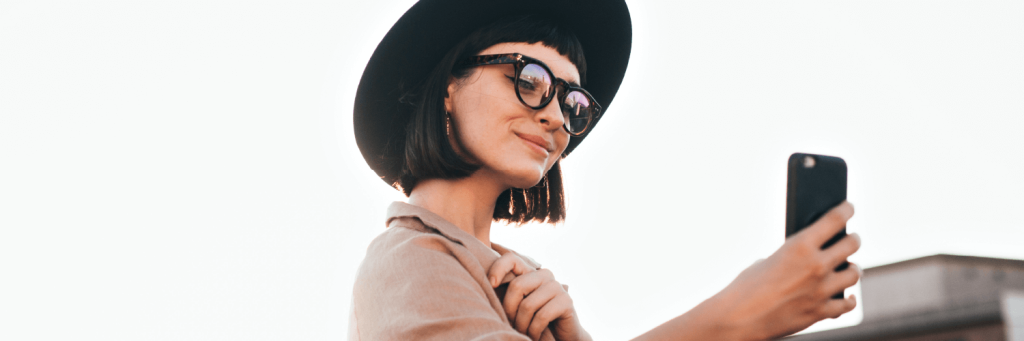 Ragazza con cappello e occhiali da vita che sorride guardando il telefono