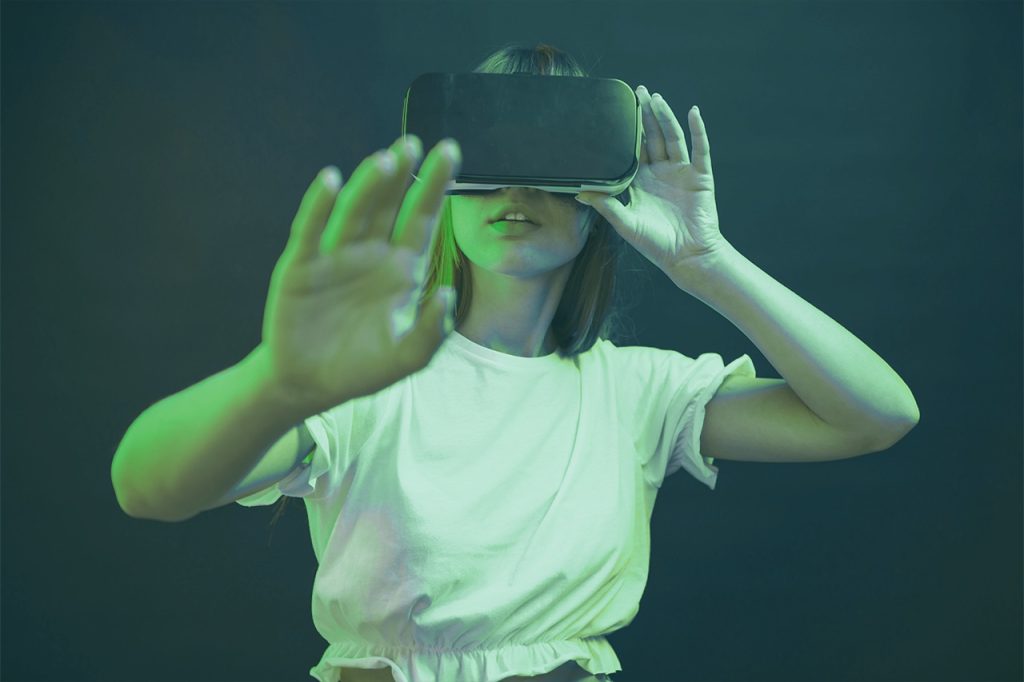 Una persona che indossa una camicia bianca utilizza un visore per la realtà virtuale, estendendo una mano in avanti come se interagisse con un ambiente virtuale, guidando la propria esperienza utente attraverso la tecnologia immersiva.
