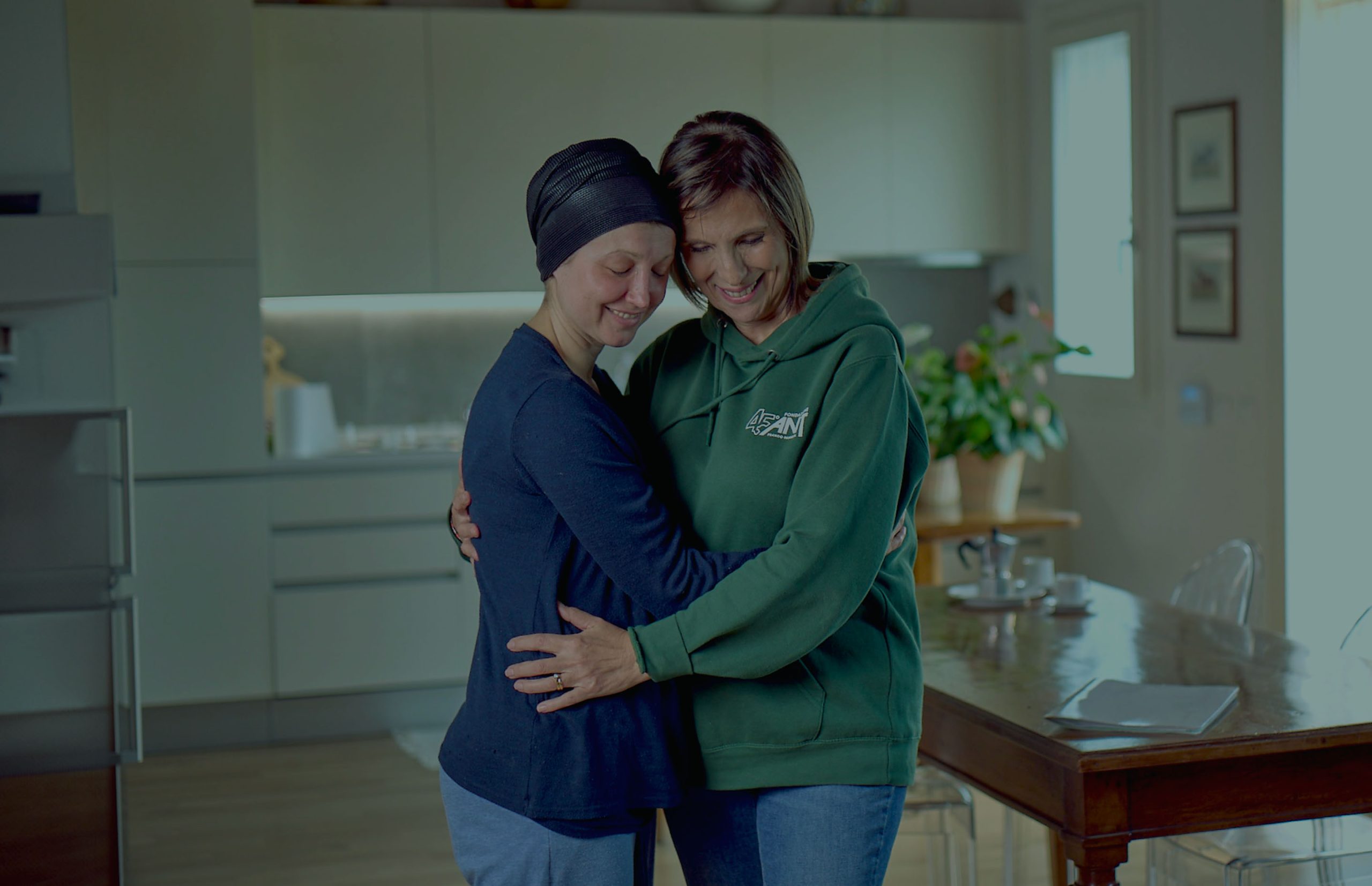 Two people hug in a kitchen—one in a beanie, the other in a green hoodie—sharing a warm moment.